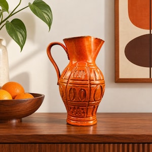 May include: A vibrant orange ceramic pitcher featuring a glossy finish and a textured, relief-patterned surface with vertical lines and oval shapes. The pitcher has a curved handle and a wide, flared rim, standing as a singular decorative vessel.