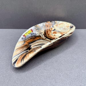 May include: A crescent-shaped tray featuring a marbled pattern of brown, black, white, and cream swirls. The surface has a glossy finish and includes a small rectangular yellow label with red borders and black text printed on the left side.