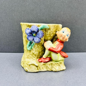 Könnte beinhalten: Keramikfigur eines sitzenden Elfen neben einer vasenförmigen Baumstumpf. Der Elf trägt eine rote Jacke und grüne Hose. Eine blaue Blume mit grünen Blättern befindet sich auf der Vase. Die Vase ist beige und hat eine strukturierte Oberfläche.