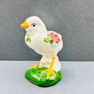 Könnte beinhalten: Eine Keramikfigur eines weißen Küken mit gelbem Schnabel und Füßen. Das Küken hat ein rosa Rosen- und grünes Blattdesign an der Seite und sitzt auf einem grünen Sockel. Die Figur ist etwa 10 cm hoch.