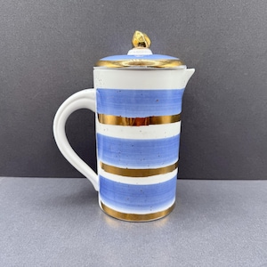 May include: A ceramic jug with a lid featuring horizontal light blue painted stripes and metallic gold bands. The jug has a white handle, a small spout, and a gold-coloured finial on top of the lid. The surface has a speckled texture.