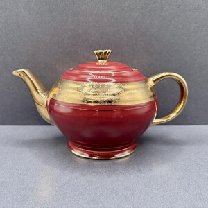 Può includere: Una teiera in ceramica rosso scuro caratterizzata da un'ampia fascia orizzontale con decorazioni in filigrana dorata attorno al centro. La teiera include un coperchio abbinato con pomello dorato, beccuccio dorato e manico dorato.