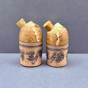 May include: Two cylindrical ceramic salt and pepper shakers with a speckled tan and brown glaze. Each shaker features a dark brown embossed wave pattern around the base and a small, angled cork stopper inserted into the top side opening.