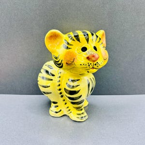 May include: A yellow ceramic tiger figurine featuring black painted stripes across its body, head, and ears. The tiger has a rounded face with large black eyes, a small pink nose, and painted whiskers, presented in a seated position.