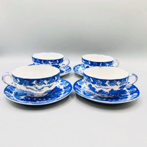 May include: Four matching teacups and saucers. The cups and saucers are white with a blue floral and landscape design. The cups have handles and the saucers are round. The set is displayed on a white surface.