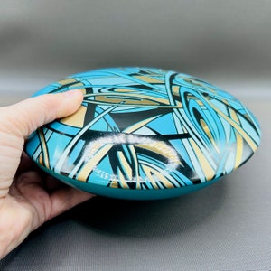 May include: A round, bowl-shaped steel tongue drum featuring a glossy finish with a complex, abstract pattern of intersecting lines and shapes in vibrant turquoise, black, and metallic gold colours, viewed from a slightly elevated side angle.