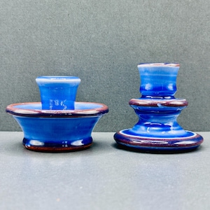Peut inclure: Deux bougeoirs en céramique avec une finition brillante, présentant une glaçure bleu vif qui passe à des tons violet foncé et marron sur les bords. Le bougeoir de gauche est large et bas, tandis que celui de droite a un design à étages.