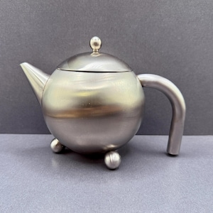 May include: A spherical stainless steel teapot with a brushed metallic finish. The teapot features a curved spout, a rounded handle, a removable lid with a small spherical knob, and three small spherical feet supporting the base of the vessel.