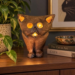 May include: A brown, textured cat-shaped lamp with a weathered finish. The sculpture features cutouts for the eyes, ears, and mouth, which emit a warm, glowing light from within. The cat is depicted in a seated position with visible paws.