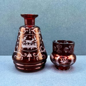 Może przedstawiać: Dwa antyczne naczynia ze szkła w kolorze rubinowej czerwieni. Większa butelka ma scenę z jeleniem i lasem wytrawioną na biało, z ozdobnymi białymi zawijasami. Mniejsze szkło ma podobny wytrawiony wzór. Oba to prawdopodobnie przedmioty dekoracyjne.