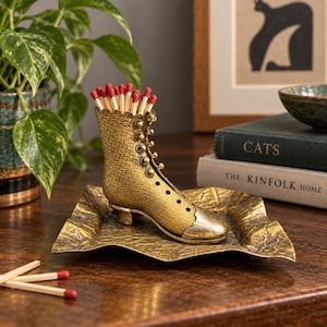 May include: A gold-toned metal boot-shaped match holder with a textured surface, featuring a row of small buttons and a smooth toe cap. The boot sits on a matching square, textured metal tray, and is filled with wooden matches with red tips.
