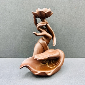 Peut inclure: Un brûleur d'encens en céramique marron représentant une main sculptée tenant une fleur de lotus au sommet, placée au-dessus d'une seconde main ouverte reposant dans une base incurvée en forme de feuille. L'objet a une finition mate uniforme.
