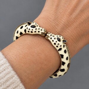 May include: A hinged cuff bracelet featuring two giraffe heads facing each other. The bracelet is finished in cream enamel with black spots and black enamel eyes, crafted with a metallic gold-tone base that highlights the giraffe features.