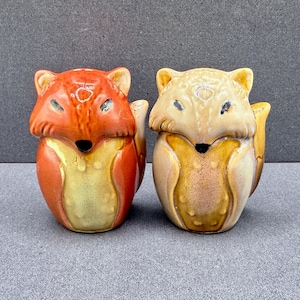Peut inclure: Deux salières en céramique en forme de renard. La salière de gauche est orange avec un poitrail crème et un nez noir. Celle de droite est de couleur beige clair et crème avec un nez noir. Les deux ont des oreilles pointues et un fini brillant.