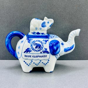 May include: A white and blue ceramic teapot shaped like an elephant, with a smaller elephant figurine on top. The teapot has a blue handle and spout, and the words "BLUE ELEPHANT" are printed on the side.