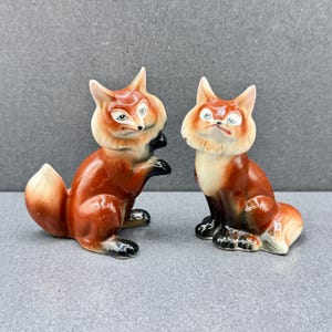 May include: Two ceramic fox ornaments. The foxes are depicted in a seated position with curled tails. They feature orange and white fur, black paws and noses. The figurines have a vintage appearance.