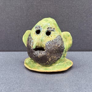 May include: A quirky ceramic face sculpture with a green head, a large black beard, and open eyes. The sculpture has a small, round base and is painted with a matte finish. The face has a surprised expression.