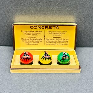 Può includere: Un set di profumi vintage con l'etichetta "CONCRETA" con tre applicatori di profumo sferici colorati in una scatola gialla. Gli applicatori sono rossi, gialli e verdi, ciascuno con un design in bianco e nero. Il testo sulla scatola descrive il prodotto.