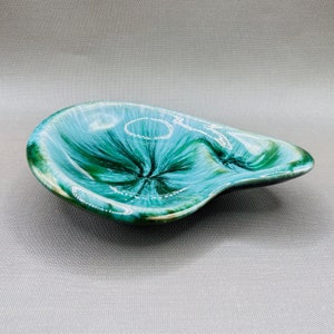 Può includere: Un piatto in ceramica a forma di rene, color turchese, con finitura lucida. Il piatto presenta un motivo a vortice di turchese scuro e chiaro, con un centro più scuro. I bordi sono verde scuro, su sfondo grigio.