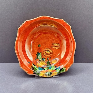 Peut inclure: Un bol en céramique à bord festonné avec un vernis orange et rouge vif. L'intérieur est orné d'un motif floral peint à la main représentant des coquelicots jaunes et orange avec des feuilles vertes, partant du fond vers le bord du bol.