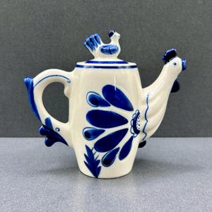 Könnte beinhalten: Eine weiße Keramik-Teekanne mit blauen Blumenmustern und einem hahnförmigen Ausguss. Der Griff und der Deckel sind ebenfalls mit blauen Akzenten versehen. Auf dem Deckel sitzt eine kleine blaue Vogelfigur. Ein dekorativer Küchenartikel.
