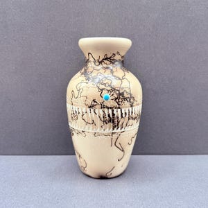 May include: A small, beige ceramic vase with black abstract line art and a turquoise accent. The vase features a decorative band with a white geometric pattern. The vase is a decorative item.