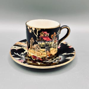 May include: A black ceramic teacup and saucer set featuring a detailed hand-painted landscape design with a red-roofed pagoda, golden trees, and a bridge. The cup has a curved handle and both pieces are finished with a thin gold-toned rim.