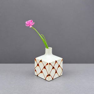 以下が含まれることがあります： 斑点のあるオフホワイトの仕上げと茶色の格子模様が施された正方形の陶器製花瓶。緑色の茎を持つ鮮やかなピンク色の花が1輪、花瓶の狭い首部分に挿されており、正方形の土台の中央から上に向かって伸びています。
