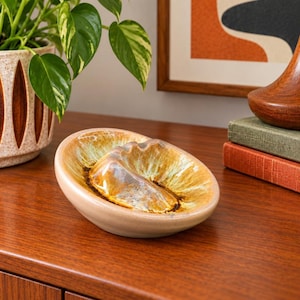 May include: A ceramic dish with a smooth, light beige exterior and a glossy, textured interior featuring a sunburst pattern of golden-brown, amber, and cream glazes. The bowl has a shallow, oval shape with a raised, moulded centre detail.