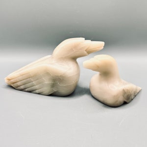 May include: Two carved stone duck figurines. The larger duck is lying down, and the smaller duck is sitting upright. Both are a creamy white colour with subtle veining, and are set against a grey background.