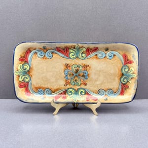 May include: Rectangular ceramic serving tray with a beige background and a blue border. The tray features a colourful floral design in red, blue, and green. The tray is supported by a small gold-coloured stand.