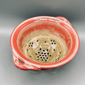 May include: A round ceramic colander featuring a vibrant red glazed rim and two side handles. The interior basin is finished in a smooth, muted beige glaze and contains numerous circular drainage holes distributed across the bottom and sides.