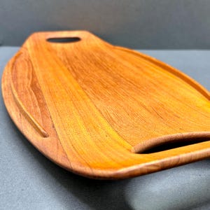 以下が含まれることがあります： 持ち手付きの木製まな板。暖かく赤みがかった茶色で、曲線的な形状。縁に溝があり、持ち手は切り抜き。木目がはっきりと見え、自然な風合いを醸し出しています。