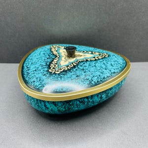 May include: A turquoise and gold trinket box with an unusual, organic shape. The lid features a gold and cream-coloured ornate design and a small, dark knob. The box has a gold rim and a textured surface.