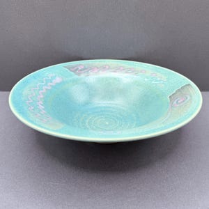 May include: A shallow ceramic bowl with a wide rim, featuring a speckled turquoise and teal glaze. The surface is decorated with faint, hand-painted light purple wavy lines and spiral patterns that follow the curve of the bowl's interior rim.
