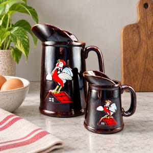 May include: Two dark brown glazed ceramic jugs featuring a raised relief design of a cockerel standing on a red roof. The larger jug has a wide spout and handle, while the smaller jug is a matching miniature version with a similar handle.