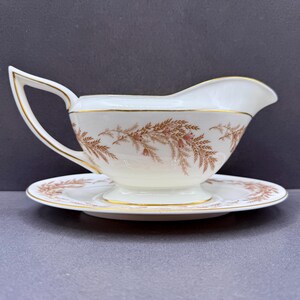 May include: A white porcelain gravy boat with an attached saucer, featuring a delicate brown fern-like botanical pattern and thin gold trim along the rim, handle, and base. The vessel has a curved handle and a wide pouring spout for serving.