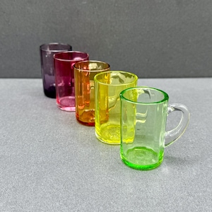 Peut inclure: Un ensemble de cinq petites tasses en verre disposées en ligne diagonale. Les tasses sont translucides et présentent un spectre de couleurs vives comprenant le violet, le magenta, l'orange, le jaune et une tasse vert clair avec une anse.