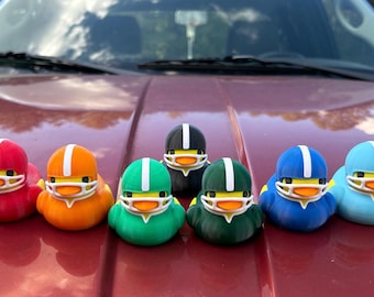 May include: A collection of colorful duck figurines, each wearing a football helmet with a white stripe. The ducks are in various colors, including red, orange, green, black, blue, and light blue. They have yellow beaks and white face masks.