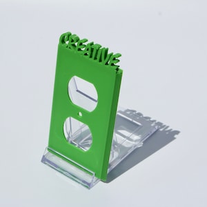 May include: A green plastic outlet cover with the word "CREATIVE" cut out in the top. The cover is on a clear plastic stand.
