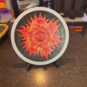 May include: A round, decorative piece featuring a vibrant, stylized sun design. The sun is rendered in shades of red and orange, with a textured appearance. It is set against a black background, framed by a gray border. The piece is displayed on a black stand.
