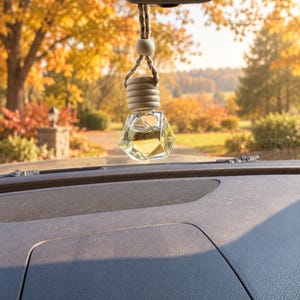 May include: A diamond-shaped glass car air freshener bottle filled with a light yellow liquid, featuring a natural wooden cap and a single wooden bead, suspended by a braided rope from a vehicle rearview mirror against a blurred autumn background.