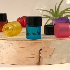 May include: A collection of small glass bottles with black lids. The bottles are filled with different colored liquids. One bottle is labeled "Lavender".