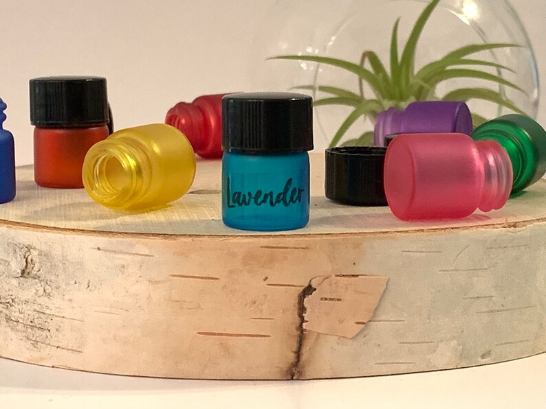 May include: A collection of small glass bottles with black lids. The bottles are filled with different colored liquids. One bottle is labeled "Lavender".