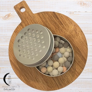 May include: A small, open, silver-colored container filled with small, round, pastel-colored balls. The container's lid, with a honeycomb pattern, is partially open. The container sits on a wooden cutting board with a handle.