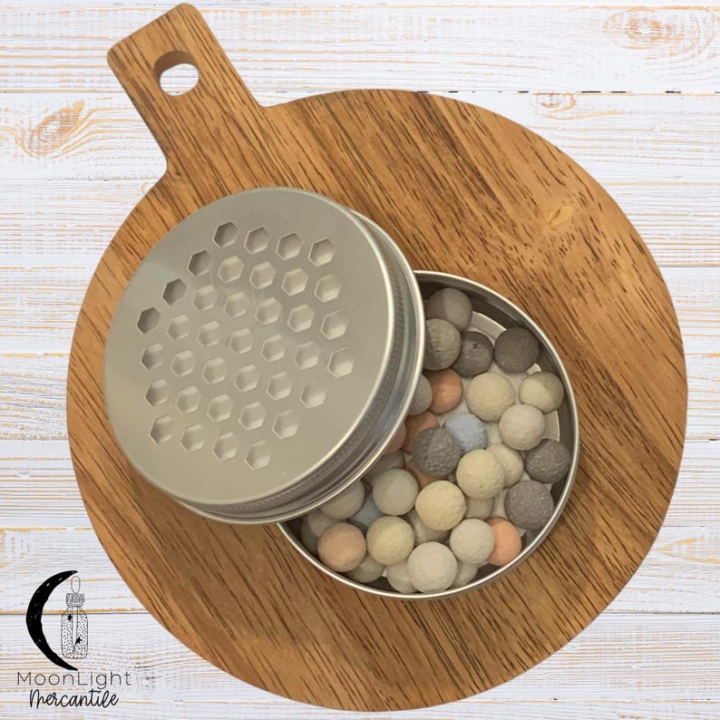 May include: A small, open, silver-colored container filled with small, round, pastel-colored balls. The container's lid, with a honeycomb pattern, is partially open. The container sits on a wooden cutting board with a handle.
