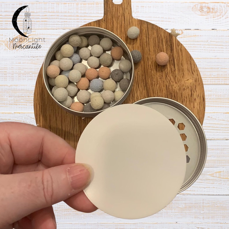 May include: A metal tin filled with small, colorful spheres, next to a wooden cutting board. The tin's lid is partially open, revealing a perforated design. A hand holds a round, off-white disc. The Moonlight Mercantile logo is in the upper left corner.