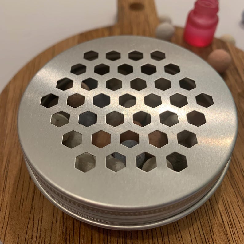 May include: A silver metal jar lid with a honeycomb pattern sits atop a jar filled with small, colorful stones. The jar is on a wooden cutting board. A small pink bottle is visible in the background.