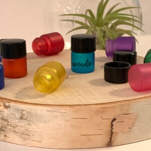 May include: Assortment of small glass bottles with black lids, filled with colorful liquids. The bottles are arranged on a wooden surface. One bottle is labeled "Lavender".