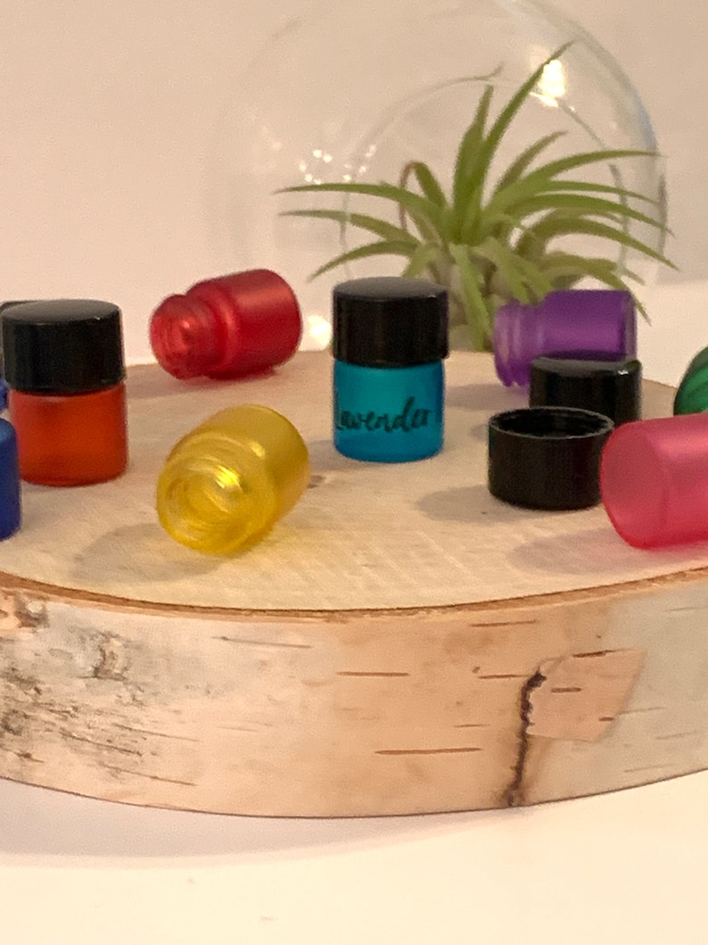 May include: Assortment of small glass bottles with black lids, filled with colorful liquids. The bottles are arranged on a wooden surface. One bottle is labeled "Lavender".